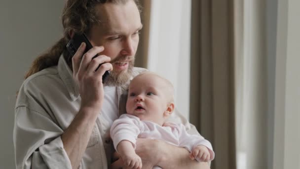 Бородатий білий чоловік батько дорослий багатозадачний тато тримає дитину вдома, розмовляє по телефону з педіатром, відповідаючи на дзвінок на замовлення з маленькою дитиною новонародженою дочкою сином відпусткою по материнській лінії — стокове відео