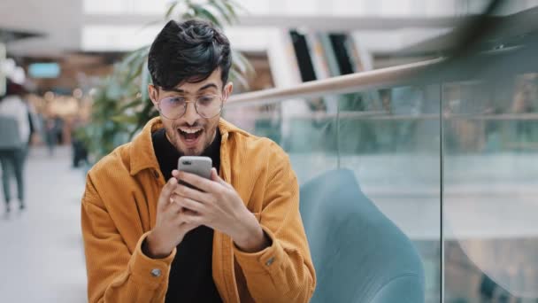 Akıllı telefon ekranına bakan genç Hintli adam beklenmedik iyi haberler alıyor gülümsüyor iyi indirimli online alışverişe hayranlık duyuyor piyangoyu kazanıyor. — Stok video
