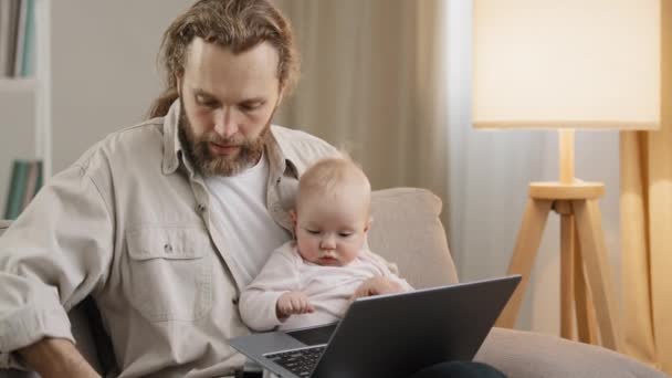 Multitasking father parent daddy business man bearded dad with little daughter son child baby infant newborn toddler working at home on sofa typing on laptop answering phone talking on mobile phone — Stock Video