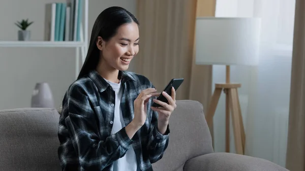 Asiática feliz chica sonriente mujer sentada sofá en casa mirando el teléfono móvil chateando navegación utilizando el servicio web wifi viendo vídeo aplicación en línea mensajes de texto con amigos teléfono inteligente ordenar compras por Internet —  Fotos de Stock