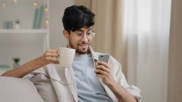 Millennial arabský chlap indický pohledný 30s muž sedí doma na gauči pití horkého čaje káva cappuccino kakao z šálku dostávají zprávu při pohledu na telefon s úsměvem chatování s přáteli s smartphonem — Stock fotografie