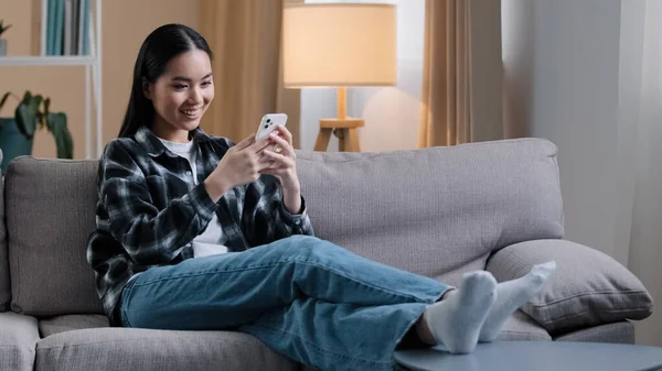 Asiatisk flicka kvinna sitter på soffan hemma avkopplande titta på nyheter foder på sociala medier välja kläder online butik internet butik dejtingsajt wi-fi smartphone shopping inlägg berättelser svepa pekskärm — Stockfoto