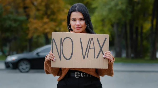 Young serious hispanic woman activist standing outdoors protesting demonstrating banner with inscription no vax shows protester cardboard sign against vaccination prohibition refusing to immunize