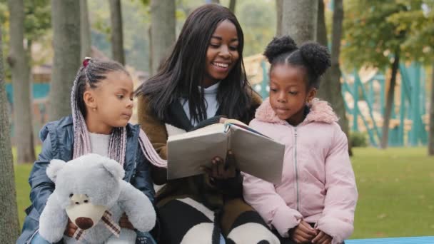 Vänlig familj sitter på bänk i parken ung mamma läser bok döttrar barn håller nallebjörn afrikansk amerikansk mor kommunicera med små flickor systrar gärna tillbringa tid med mamma utomhus — Stockvideo