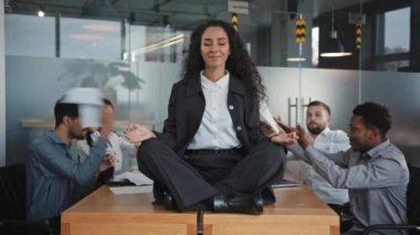 Genç İspanyol kız Lotus pozisyonunda masada oturuyor. Sakin, uyumlu iş kadını meditasyon yapıyor. Stres için yoga yapan sinir bozucu meslektaşlarını görmezden geliyor.