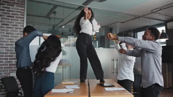 Radostné rozmanité zaměstnance oslavující dokončení vítězného projektu vedoucí žena s úsměvem šťastně tančí na stole multiraciální kolegové na kancelářské party šťastný přátelský pracovní skupina baví společně — Stock video