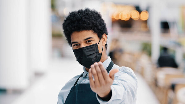Friendly african masked guy male waiter salesman man in medical face protective mask restaurant cafe kitchen bar worker pointing hey you at camera welcoming gesture come here approach welcome inviting