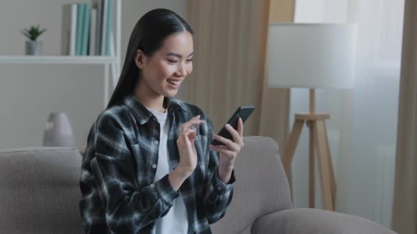 Азійська щаслива жінка, яка сидить вдома на дивані і дивиться на телефонні розмови за допомогою Wifi веб-сервісу перегляду відео смс з друзями смартфони замовлення інтернет-магазинів — стокове відео
