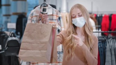 Tıbbi maske takan neşeli kadın müşteriler, alışverişlerle birlikte mutlu kızlar, mağazada baş parmak gösterip dans ediyorlar. Başarılı pazarlığın tadını çıkarıyorlar. Karantina sırasında güvenli alışverişi onaylıyorlar.