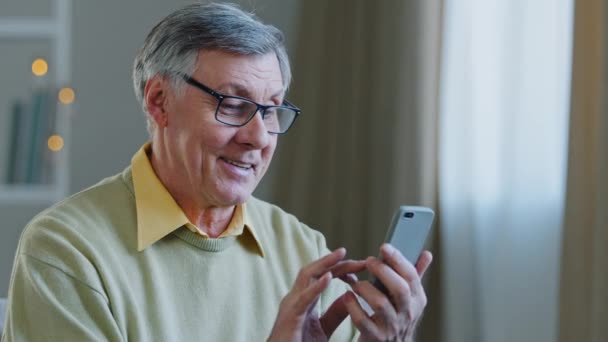 Heyecanlı orta yaşlı, gözlüklü, elinde telefon olan emekli adam internet uygulaması kullanan arkadaşlarıyla veya çocuklarıyla online alışveriş iletişimi kuruyor. Büyükbabam başarılı bir şekilde modern teknoloji öğreniyor. — Stok video