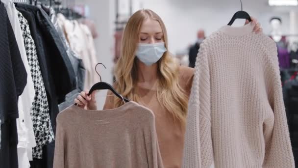 医療マスクの若い女の子は、ストアで服を選択女性消費者は、 2つのセーターの間で選択すると、新しいファッショナブルなイメージを収集します。 — ストック動画