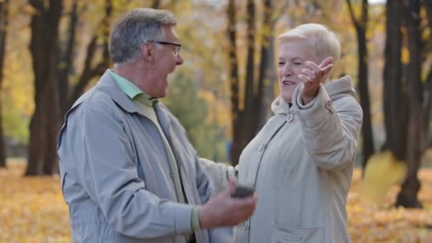 Sonbahar parkında neşeli yaşlı kadın ve adam ellerinde telefon tutan yaşlı çift zaferi kutlayan iyi haberler aldılar. Mutlu emekliler gülüyor. Açık havada sarılıyorlar. — Stok video