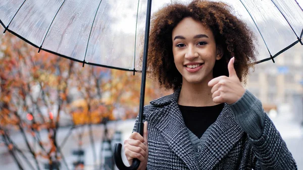 Afryki szczęśliwy sukces dziewczyna kręcone wielorasowy kobieta trzyma przezroczysty parasol pozowanie w jesiennym mieście w deszczu patrząc na aparat pokazuje kciuk w górę zgadzam się rekomendacja gest zadowolony z pogody — Zdjęcie stockowe