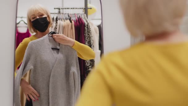 Vit medelålders kvinna kund kund i skyddande mask i butik kläder butik står i omklädningsrum tittar i spegel poserar plockar upp outfit väljer kläder mode utseende shopping koncept — Stockvideo