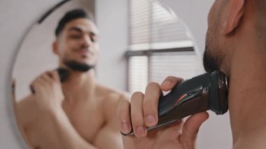 Ayna yansıması, yakışıklı Hintli Arap Kafkas sakallı adam sakalını tıraş ediyor, tıraş makinesiyle tıraş ediyor sabah banyoda tıraş oluyor, güzellik rutin erkek yüz bakımını banyoda yapıyor.
