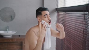 Mutlu dans eden, beyaz sabunlu, sakallı, yakışıklı ve yakışıklı Arap adam banyodaki aktif erkek banyoda tıraş olurken, traş olurken, evde dans ederken, eğlenceli, hijyen ritüelinde şarkı söylüyor.