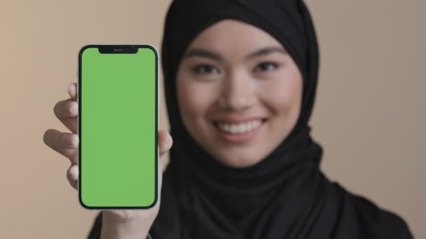 Ritratto ragazza asiatica musulmana donna islamica in hijab nero sorridente mostrando telefono cellulare con schermo verde che tiene smartphone guardando fotocamera promo video gadget pubblicità dispositivo telefono online — Video Stock