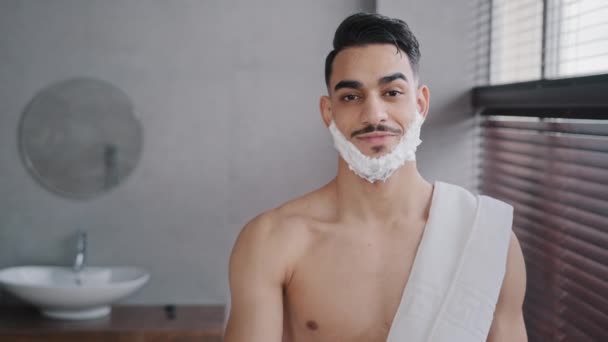 Divertente ispanico indiano arabo nudo uomo 30s barbuto ragazzo in piedi in bagno a casa con schiuma bianca saponata sulla barba gel da barba sorridente mostrando rasoio guardando la fotocamera si prepara a radersi cura del viso — Video Stock