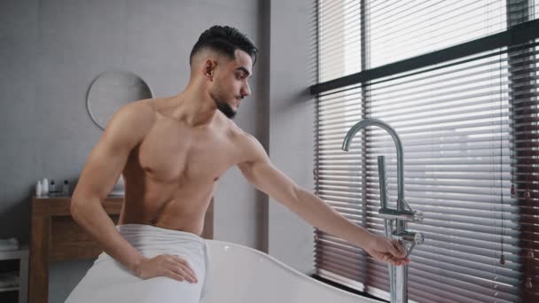 Millennial sexy nackte muskulöse indische arabische Mann Kerl mit Bart Handtuch um Hüften gewickelt sitzt auf Badewanne im Badezimmer zu Hause drehen Wasserhahn Verkostung heißes kaltes Wasser mit der Hand immer bereit zum Baden — Stockvideo