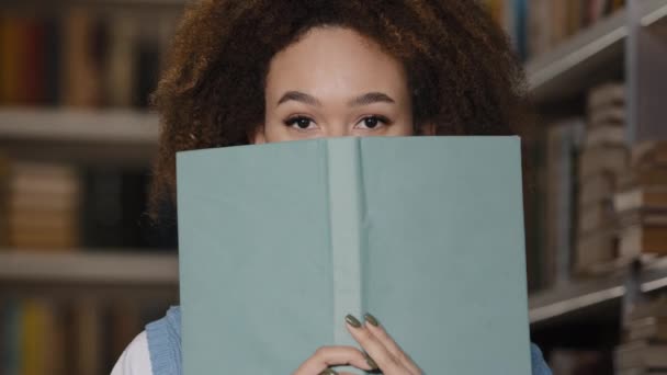 Close-up jong gelukkig speels meisje student verschuilen achter boek gluren uit voor leerboek glimlachen tandachtig glimlach vreugde spelen flirten lachen kijken naar camera genieten van lezen in de universiteitsbibliotheek — Stockvideo