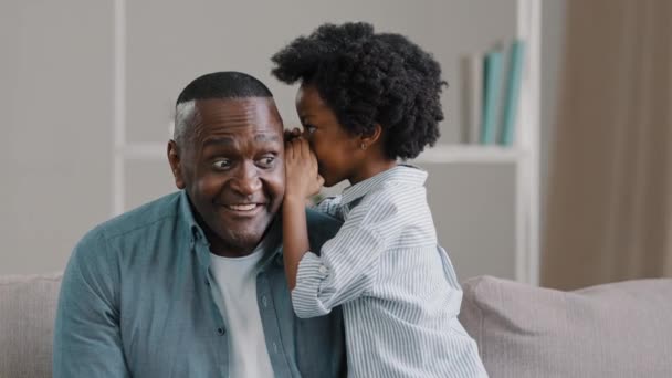 Lille afrikansk amerikansk pige deler hemmelighed med far fortæller fantastisk sjov historie i øret sjov far smilende lytter til sød datter sidder på sofaen tillidsfuldt forhold nærhed forældre og barn – Stock-video