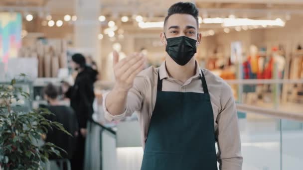 Wesoły latynoski kelner zaprasza gości szczęśliwy sprzedawca w geście fartuch powitanie klientów młody arabski właściciel małej kawiarni biznesowej przyjazny pracownik uśmiech w masce medycznej patrząc na aparat — Wideo stockowe
