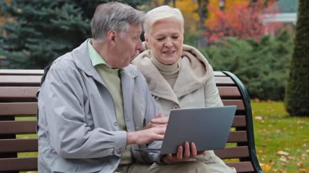 Mutlu kaygısız yaşlı büyükanne ve büyükbaba sonbahar parkındaki bankta oturup dizüstü bilgisayarda birlikte yazarak internet haberlerini tartışarak olgun çiftlerin online sipariş vermesini kolaylaştırıyor modern teknoloji kavramını kullanıyoruz. — Stok video