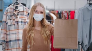 Kapalı giyim mağazası onaylı pandemik standı sırasında tıbbi maskeli mutlu genç bir kadın alışveriş torbaları gösteren bir kız jest tüketicisi düşük fiyatlı bayan indirimi gösteriyor.