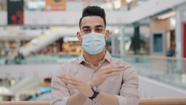 Молодой латиноамериканец в медицинской маске показывает запрещающий жест серьезный араб мужчина в респираторе стоять в торговом центре запрет во время коронавирусной пандемии ограничение посещать общественные места предупреждение эпидемии опасности — стоковое видео