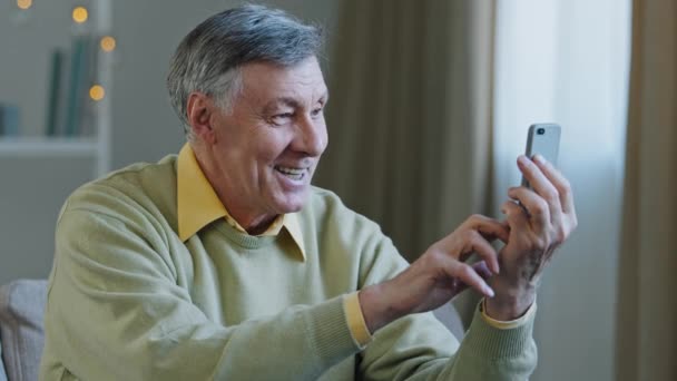 Счастливый пожилой пожилой мужчина держит телефон сделать видеозвонок улыбается дедушка глядя на экран смартфона удаленной связи использовать современные устройства положительные старые мужские говорить по телефону поделиться хорошей новостью — стоковое видео