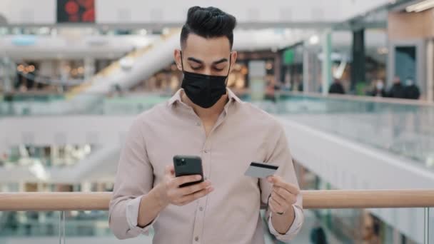 Hombre joven árabe serio en la máscara de la cara médica se encuentra en el centro comercial celebración de uso de la tarjeta de crédito aplicación de teléfono inteligente hacer el pago comprar tipo hispano haciendo orden en línea reserva indio macho contento fácil transferencia rápida de dinero — Vídeos de Stock