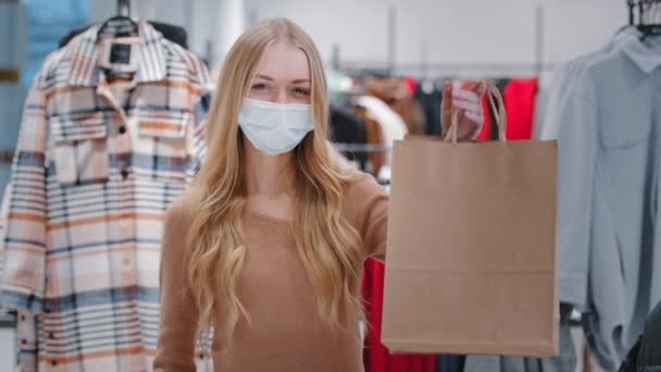 Närbild glad ung kvinna i medicinsk mask under pandemi stå i kläder butik godkännande gest flicka konsument visar shoppingpåsar pekar finger dam nöjda låga priser rabatt visa tummen upp — Stockvideo