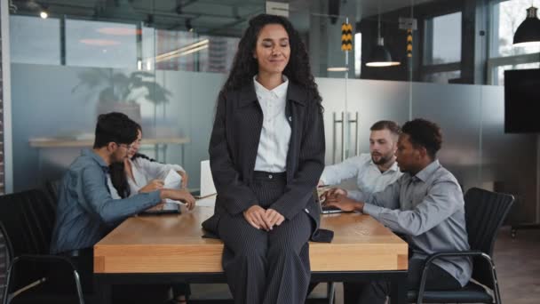 Tevreden glimlachende jonge Spaanse zakenvrouw zit achteloos op tafel in bestuurskamer multiraciale werknemers op de achtergrond bespreken werk project kantoormedewerkers brainstormen succesvolle meisje voorkomen rush — Stockvideo