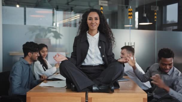 Arbete kaos kontor multiracial team i bakgrunden ung balanserad latinamerikansk flicka i Lotus position sitta på bordet psykisk hälsa kvinna ler fredligt lugn och balans undvika konflikt inre harmoni — Stockvideo