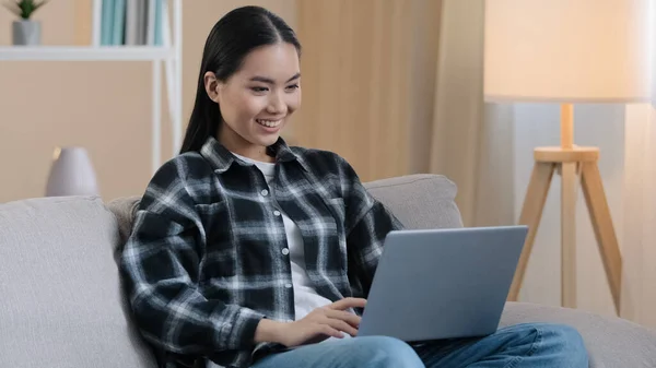 Smiling asian woman happy student girl freelancer sitting on couch using laptop typing message email lady chatting on computer browsing internet media at home easy e-learning studying working online