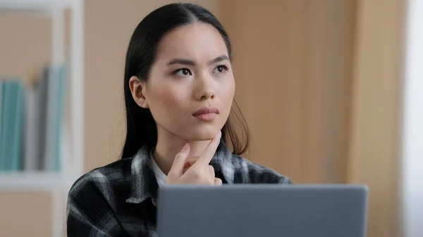 Penser fille étudiante asiatique coûteux portrait de femme se trouve en face de l'ordinateur portable faire tâche en ligne à la recherche d'une solution au problème se sent douteux profondément dans les pensées prend la décision vient avec idée trouver réponse — Photo