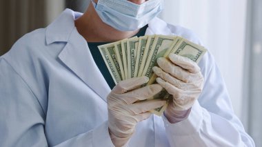 Yakın plan tanınmayan doktor hemşire, doktor, cerrah, beyaz önlüklü, koruyucu yüz maskeli, lateks eldivenli, yığınla para tutan banknotlar, klinikte rüşvet alan banknotlar.