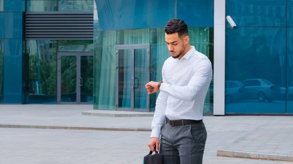 Angry sad upset hispanic business man bearded male client lawyer salesman with laptop case wears formal shirt looks at wrist watch worried about late time rushing for meeting or taxi standing outdoors — Stock Photo, Image