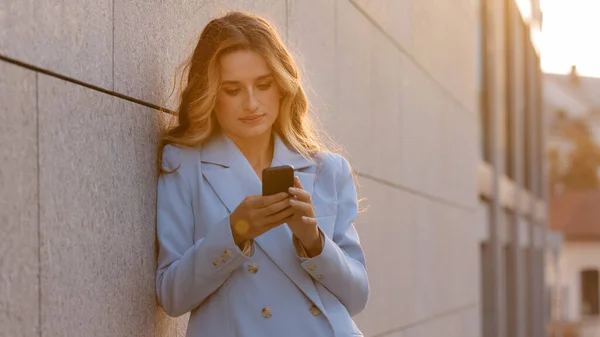 Belle caucasienne souriante heureuse femme d'affaires debout près du mur sur la rue en arrière-plan de soleil regardant dans le téléphone mobile chat smartphone acheter en ligne dactylographie message nouvelles en utilisant l'application — Photo