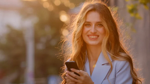 Piękna młoda dziewczyna kobieta stoi na ulicy na zewnątrz w promieniach słonecznych o zachodzie słońca patrząc na aparat gospodarstwa telefon komórkowy wiadomość online oglądanie wideo ze smartfonem na czacie korzystających internet wifi — Zdjęcie stockowe