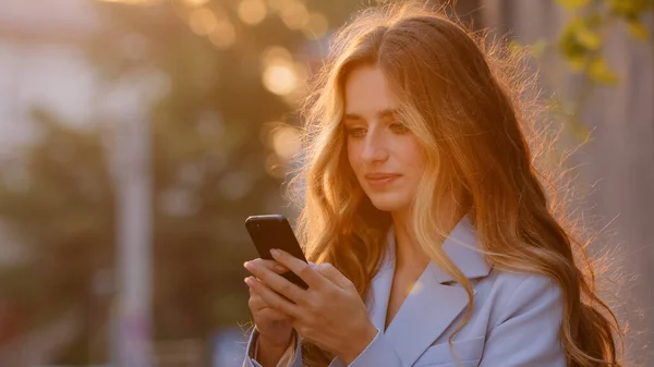 Piękna młoda dziewczyna kobieta stoi na ulicy na zewnątrz w promieniach słonecznych o zachodzie słońca patrząc na telefon komórkowy odpowiadając wiadomość online oglądanie wideo ze smartfonem na czacie korzystających internet wifi — Zdjęcie stockowe