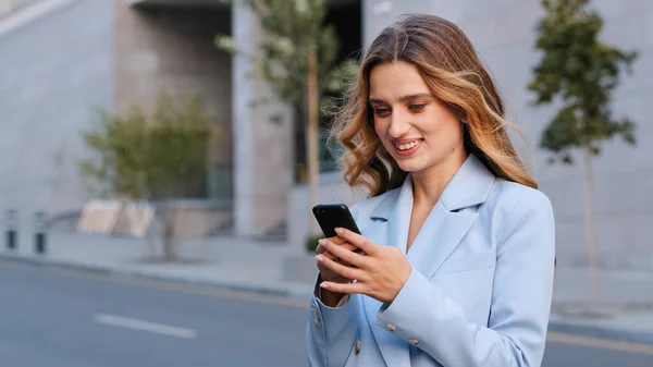 Portrait féminin en ville jeune fille blonde femme d'affaires utilisateur se tient dans la rue en regardant téléphone mobile appareil sans fil gadget en utilisant smartphone pour discuter en réseau lecture en ligne nouvelles sourire heureux — Photo