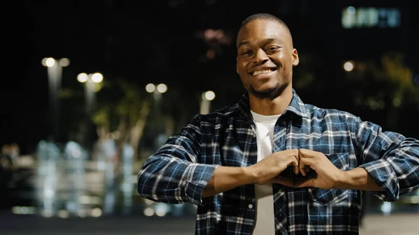 夜の街に立つアフリカ系アメリカ人の優しい男は、カメラを見て愛情のあるロマンチックな肖像画は指で心の形を作る。手で優しさのサポートを示す素敵な男の子 — ストック写真