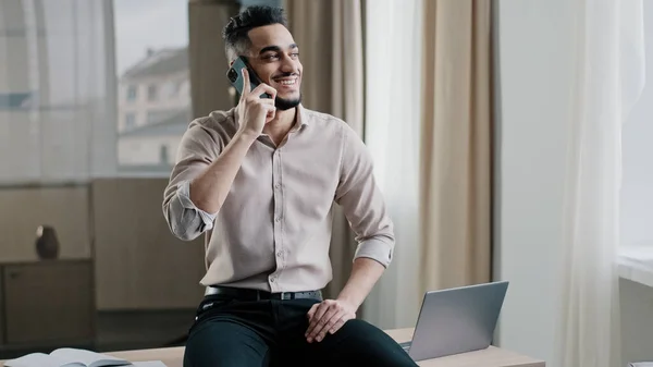 Улыбающийся араб расслабленный предприниматель-мужчина, сидящий на столе в рабочем месте, проводит переговоры удаленный телефонный разговор с клиентом, обсуждая с коллегой по мобильному телефону — стоковое фото