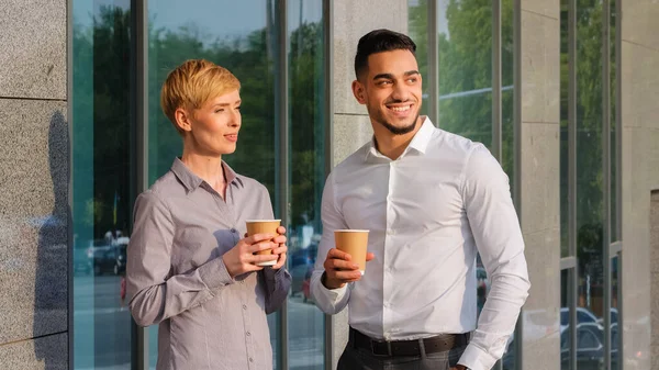 İki çok ırklı iş ortağı çok ırklı iş arkadaşı Arap erkek ve beyaz kadın şirket binasının yanında durmuş karton bardaklardan kahve içerek iş arasında mola veriyorlar. — Stok fotoğraf