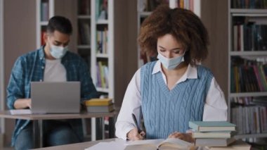 Tıp maskeli genç kız kütüphanede oturmuş not defterine not yazmaya hazırlanıyor. Kameraya bakıyor ve dizüstü bilgisayar öğrencilerinin ders çalışmalarına çalışan işaret fişekli adamı gösteriyor.