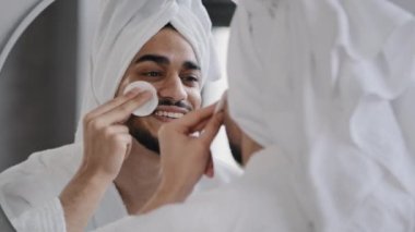 Komik, yakışıklı, sakallı, beyaz banyo havlusuyla Arap sakallı adam aynadaki yansımaya bakıyor. Güzel görünümlü erkek, evde temizlik yaparken pamuk peçeli güzellik prosedürlerini kullanıyor.