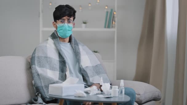 Sick arab man in medical face protective mask sitting on couch at home ill guy wrapped in plaid looking at camera with sadness suffering seasonal virus covid coronavirus pandemic contagious infection — Stock Video