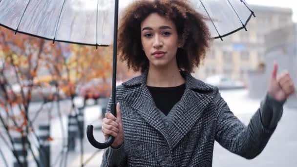 Afrykański amerykański smutny dziewczyna kobieta z kręconymi włosami z przezroczysty parasol pokazuje gest dezaprobaty odmowa sprzeciw kciuk w dół negatywna reakcja brak odpowiedzi stoi w mieście w deszczu zła pogoda — Wideo stockowe