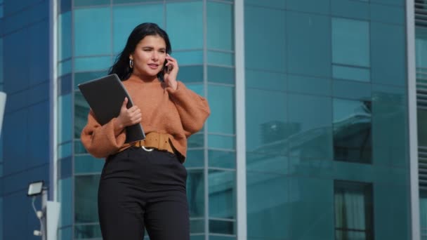 Happy businesswoman stands on background office building speaks on mobile phone answers friendly call communicates with friend using smartphone spends free time outdoors holding folder with documents — Stock Video